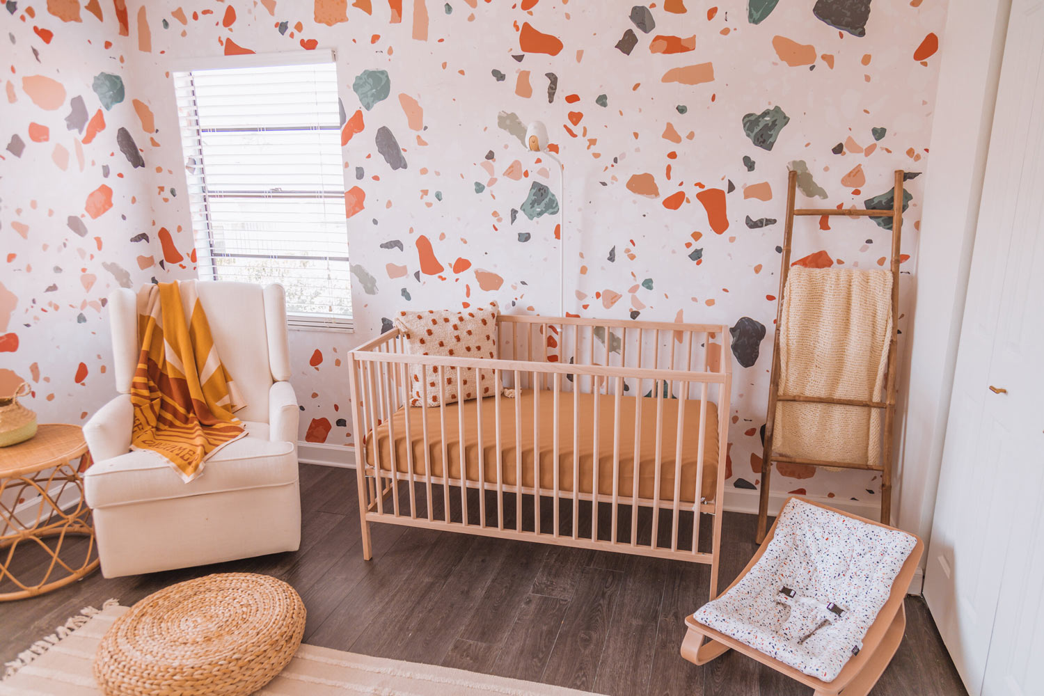 Warm tone gender neutral baby nursery interior room, with light wood ikea crib, livettes terrazzo removable wallpaper, eclectic bohemian interior style