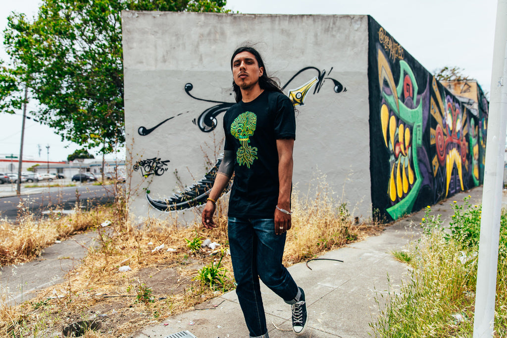 Guy in front of colorful mural wearing the Ancient Roots skull and roots design on black tee.