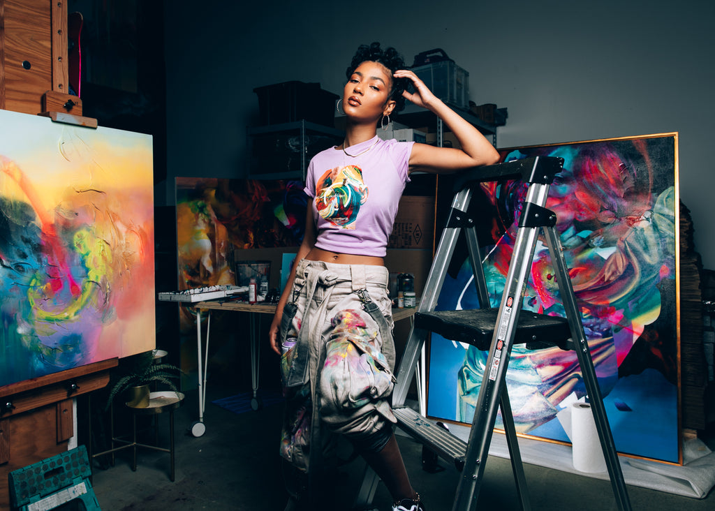 Female model in Hueman's studio wearing one of the collab tees, next to one of Hueman's paintings.
