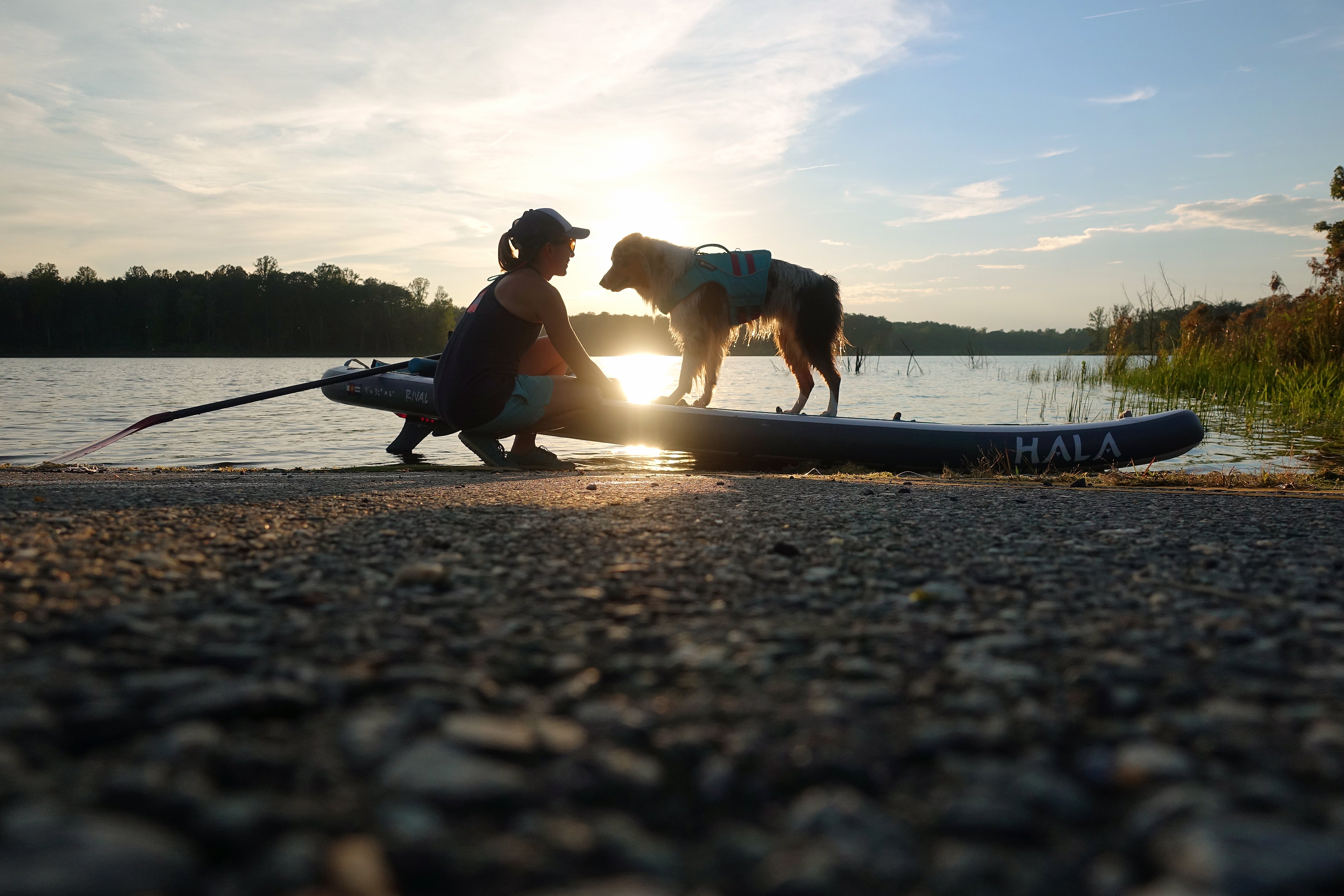 Bodie in a Float Coat stands on paddleboard on shore while Maria holds the board.