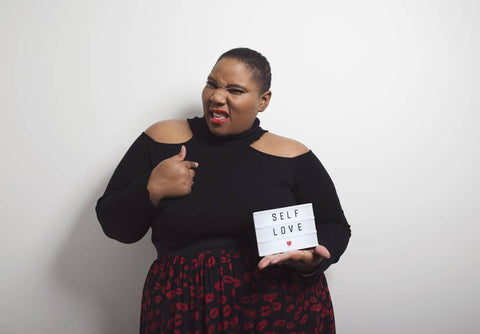 Plus-size model holding 'self love' lightbox and smirking against white wall.