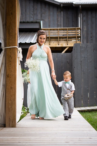 A bridesmaid walks down the aisle in a Mint Green wedding dress, the Henkaa Sakura Maxi Convertible Dress, holding Ashley's son's hand.