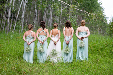 Ashley and her bridesmaids stand in a field holding Baby's-breath bouquets behind their backs. Bridesmaids are wearing Henkaa Convertible Dresses in Mint Green.