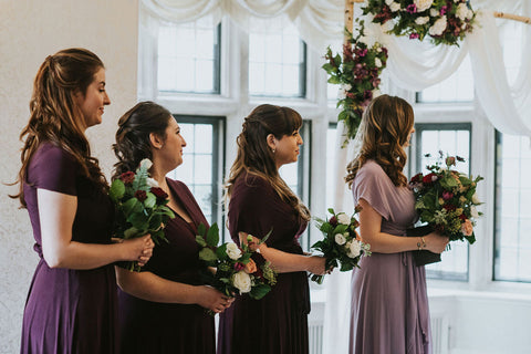 Averie's three bridesmaids and maid of honour stand holding bouquets during the wedding ceremony. Bridesmaids are wearing Henkaa Sakura Maxi Convertible Dresses in Eggplant Purple and Dusty Purple.