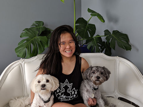 Sam sits on a couch with her two dogs Dallas and Shep, a Monstera houseplant behind them.