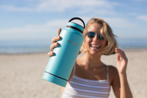A woman holds up a blue insulated water bottle on the beach