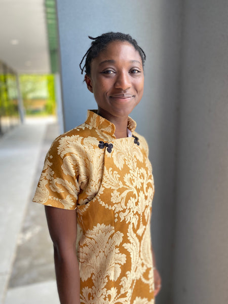 Woman standing outside, smiling, wearing a gold brocade Cheongsam