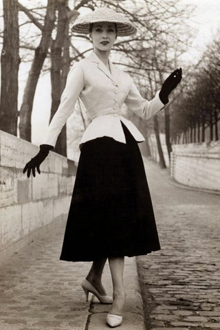 black and white image of a woman wearing a dark skirt, light top, and a hat and gloves.  Suit is the 1950s New Look.