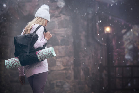 Blonde girl wearing a white beanie and a pink long sleeve walks outside in a wintery atmosphere carrying a black backpack holding a yoga mat
