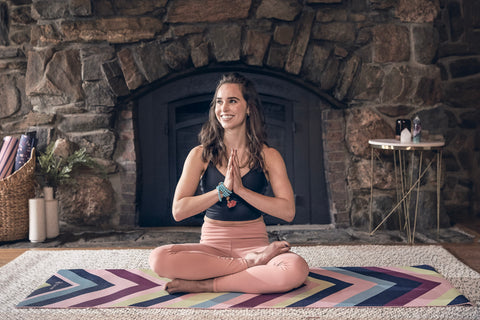 A dark haired girl wearing a black yoga top and peach yoga bottoms sits smiling on her yoga mat with her hands at her heart in front of a stone fireplace