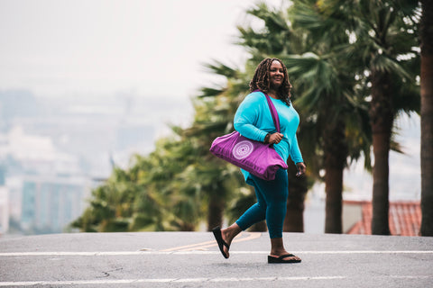 woman walks across palm tree lined street with yoga mat bag