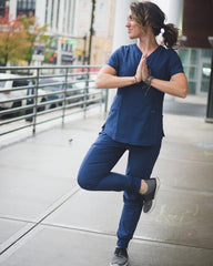 a brunette nurse with a stethoscope and scrubs stands outside in a city doing tree pose