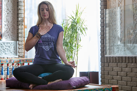 blonde girl in yoga attire sits in meditation cushions while placing her hand on her heart