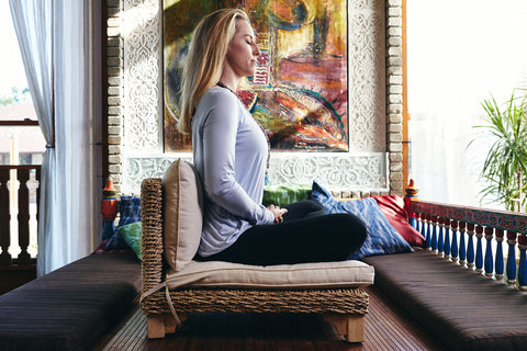 blonde girl wearing a long sleeved shirt sits meditating on a low chair in fancy room