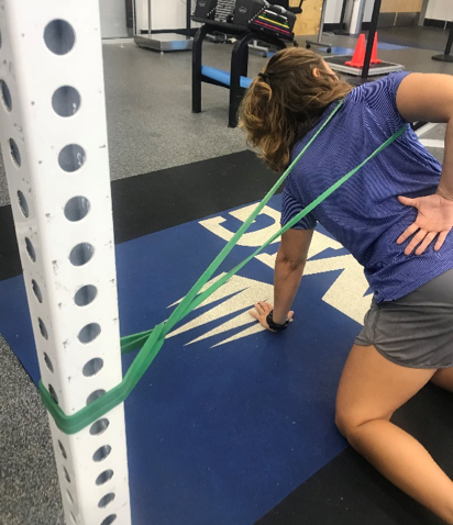 woman works out at a gym and uses a green exercise band to stretch her right shoulder 