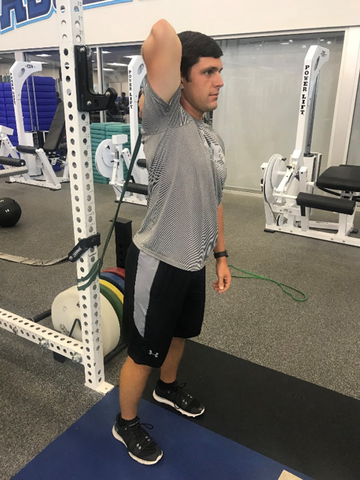 Man in a uses a long green SPRI superband to stretch his shoulder at a gym