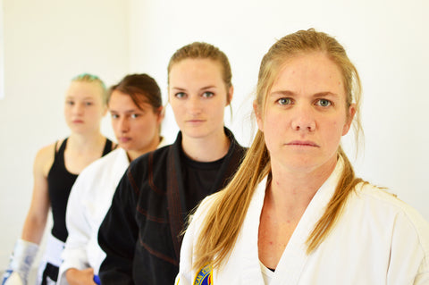 Four women standing in a line one in front of the other with serious looks on their faces. Each dressed in different martial arts uniforms. 