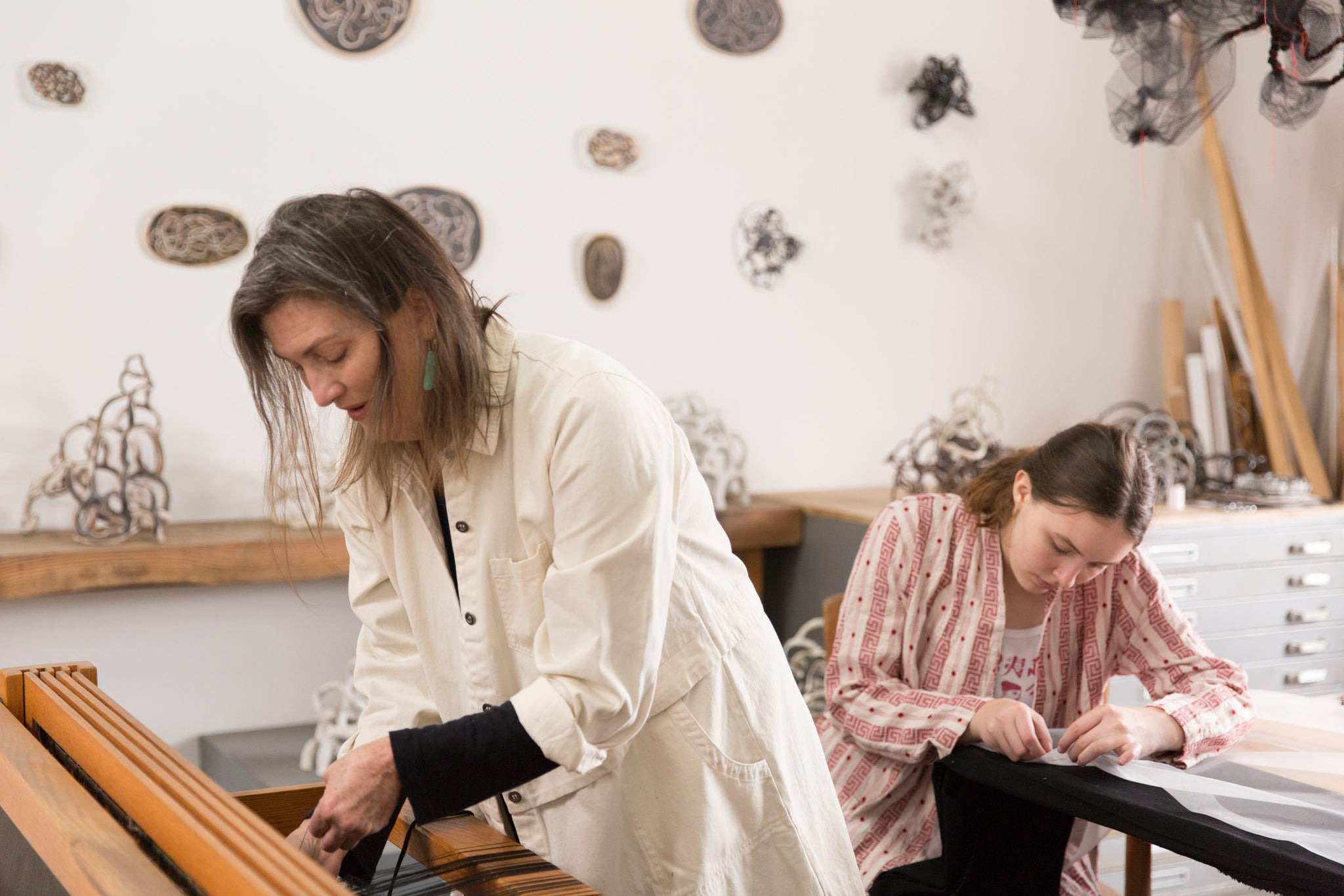 artist Laura Cooper working at loom in foreground with her daughter work at table in background