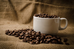 Cup of coffee on a burlap sack surrounded by roasted coffee beans.