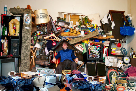 A man sitting in a chair amongst pile of stuff that surrounds him on all sides, even threatening to fall on his head.