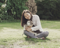 A girl holds her small beagle dog in her lap surrounded by green grass and trees. The girl is smiling at the camera while her dog looks off to the side.