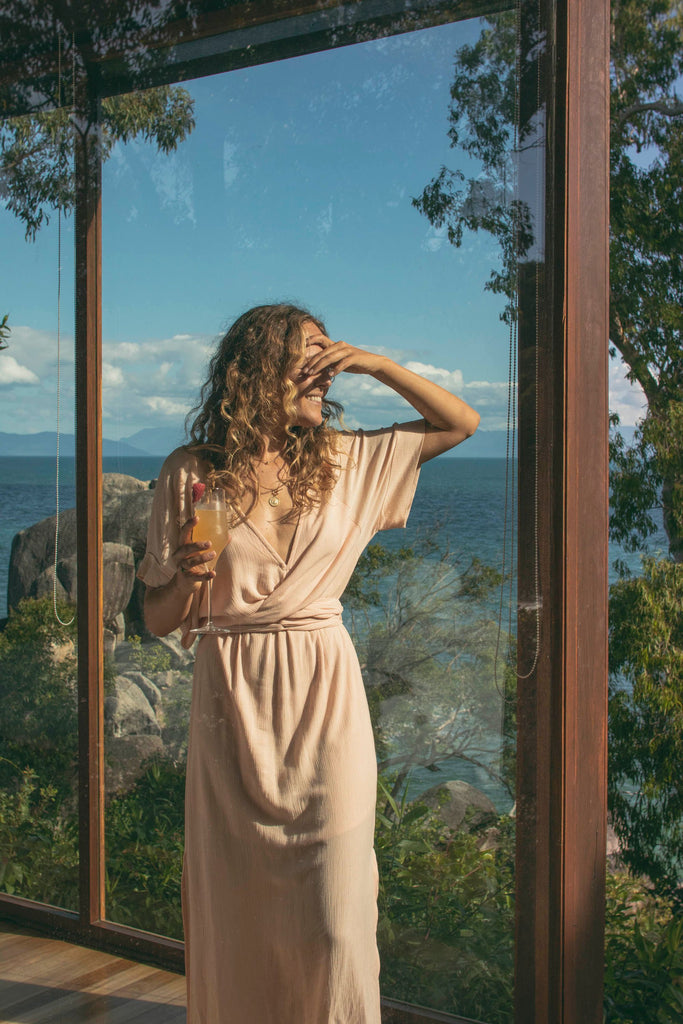A woman wearing light pink wrap dress standing in front of a window covering her face while holding a drink