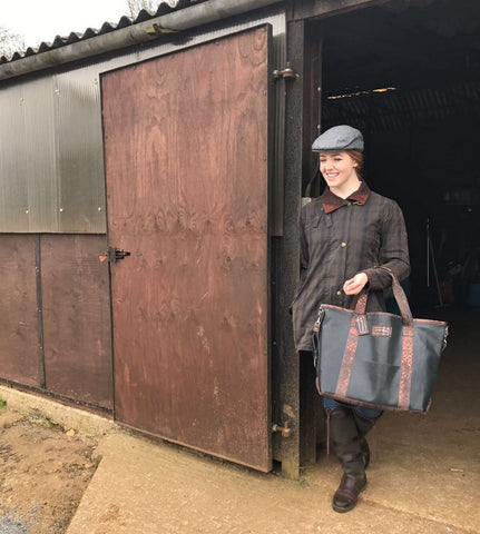 Harriet poses outside a barn in a barber jacket with jeans, high boots and a flat cap…paired with our Richmond Tote.