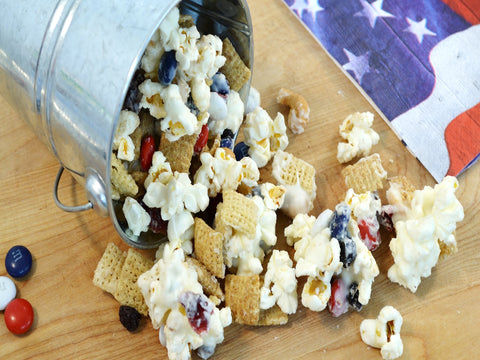 All-American Snack Mix Recipe spilling out of a metal bucket with an American flag painting to the right of the popcorn