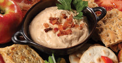 A beige crockpot of RADA's savory Applewood Smoked Bacon Dip surrounded by an array of assorted crackers and topped with a garnish and bacon bits