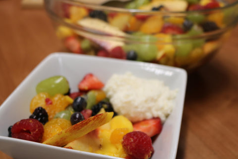 A colorful bowl of fruit salad with peaches, raspberries and apple slices in a white bowl next to a larger glass bowl of mixed fruit