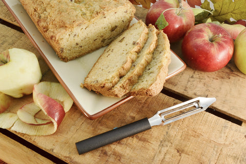 A RADA high carbon stainless steel Vegetable Peeler with a black resin handle next to a sliced loaf of banana bread and several peeled and unpeeled apples