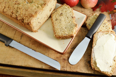 A RADA high carbon stainless steel Party Spreader and 6" Bread Knife with black resin handles next to a loaf of sliced and buttered bread