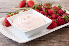 A white bowl of our white chocolate raspberry dip on a plate full of bright strawberries on top of a white tablecloth