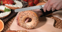 A RADA Bagel Knife slicing a whole grain bagel in half on top of a wooden cutting board