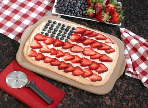 An American flag colored fruit pizza with sliced red strawberries and blueberries next to a RADA Cutlery Pizza Cutter and a couple handkerchiefs