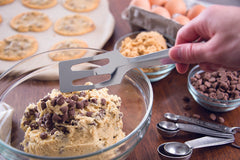 A Rada Serverspoon hovering above a glass bowl of cookie batter with a few measuring spoons and another smaller glass bowl of chocolate chips