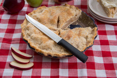 A RADA Slicer knife with a black resin handle with a black resin handle on top of a apple pie with a few apple slices beside the pie over a red checkered table cloth