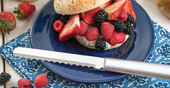 A RADA Bagel Knife with a silver brushed aluminum handle next to a bagel stuffed with raspberries, blackberries and sliced strawberries on top of a purple plate over a blue placemat
