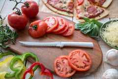 A RADA Cutlery stainless steel Tomato Slicer on a brown cutting board with slices of red tomatoes and peppers around the cutting board with a bowl of cheese to the right and a pizza farther up on the table