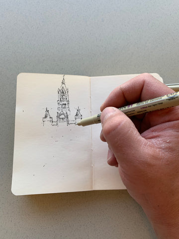 Image of a hand being supported by one side of a sketchbook, while drawing a building on the opposite page