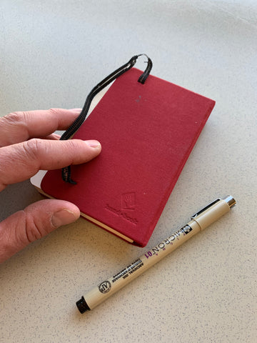 Image of a mans left hand holding the corner of a small, red travel sketchbook