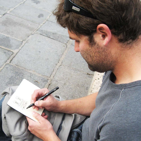 Image of a man (Eric Jacoby) sitting on a low concrete wall with a blazer in his lap, sketching Notre Dame Paris in a small travel sketchbook