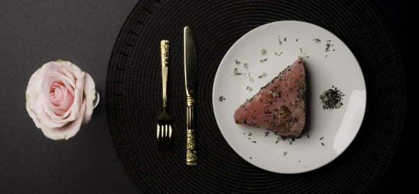 Pink rose beside a plate with a pink tuna steak and lavender and green tea salt, black background