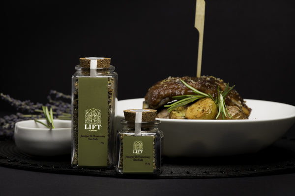 Large and small bottles of Lift Juniper and Rosemary Salt in front of a plate of pork ribs and roasted potatoes, from the side