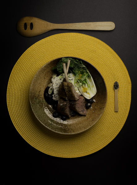 Lamb chops, rice, and bok choy with Lift Lavender and Green Tea salt, on a yellow round placemat, beside a slotted wooden spoon, from above