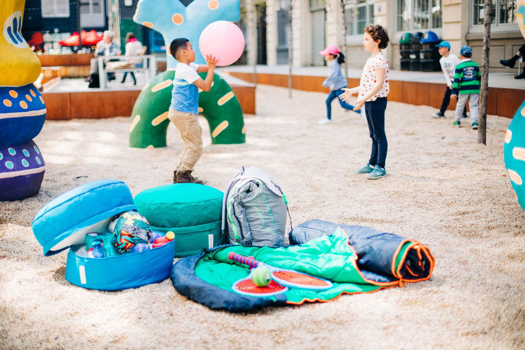 Two children playing outdoors with a mimish sleep-n-pack on the floor and a pouf