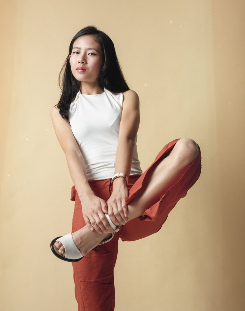 Elisa dressed in Gene Ave's sleeveless white top and long orange pants. She is posing with both hands holding her leg up by the ankles while balancing on the other leg..