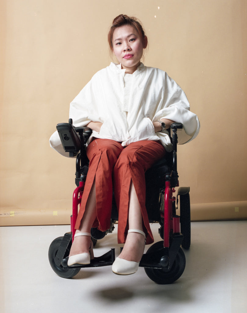 Yean Cheng seated in her wheelchair. She is wearing long orange pants and textured white cape from the Gene Ave collection.