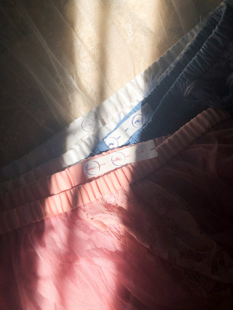 Pink, blue and cream coloured skirts layered on top of each other, with adjustable buttons and waistband exposed. A strip of sunlight shining in the middle and on the buttons.