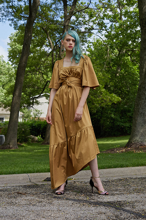 Cynthia Rowley 2019 Resort Collection features a saffron colored asymmetrical maxi dress belted at the high waist, and finished with gold and blush colored heels.